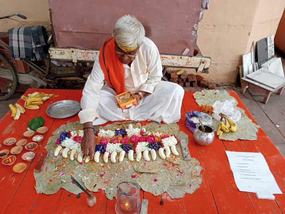 Brahmin pandit performing Online Pind Daan in Varanasi with 16 rice ball pinds arranged on banana leaves with marigold and rose flowers, lit diyas, and a handwritten list of departed souls names and gotra placed beside the ritual setup on a red wooden platform